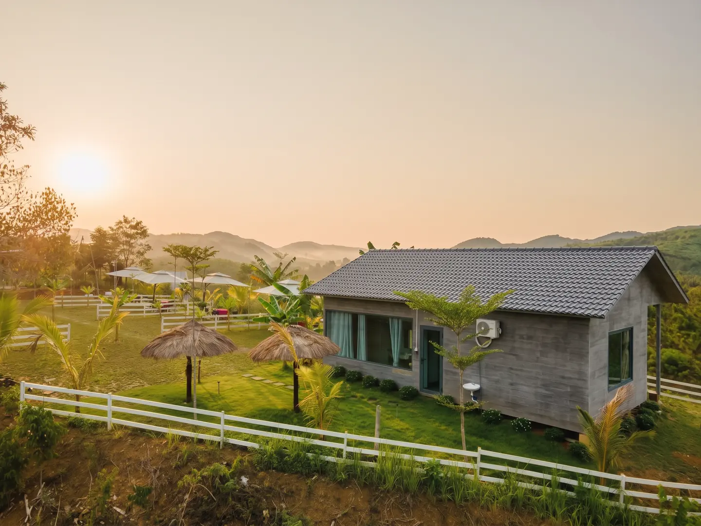 Bungalow mái ngói xám, mặt kính lớn, sân cỏ rào trắng và hai chòi tranh trên đồi lúc bình minh